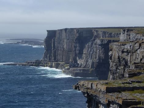 Høje kalkstensklipper langs Irlands vestkyst med lagdelte klippeformationer. Hvide bølger bryder mod de mørke klipper.