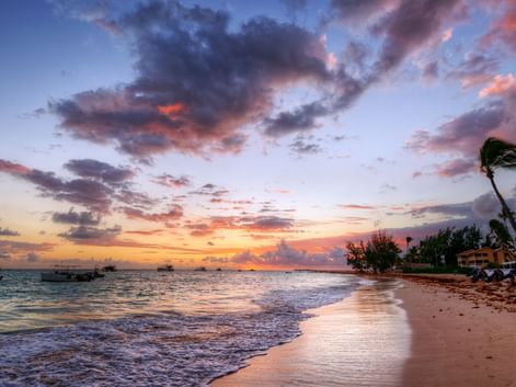 Dramatisk solnedgang over en strand på Bali med palmer i vinden, fargerike skyer reflektert på våt sand og båter utenfor kysten.