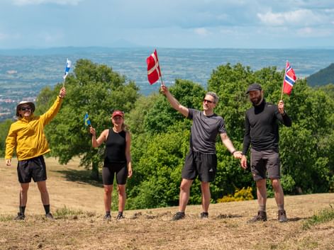 Four hikers holding flags from different countries on a hilltop along Via Francigena near Orvieto, with green hills and valleys in background.