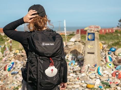 Rear view of pilgrim with backpack and scallop shell on Portuguese Camino, walking past stone markers with yellow arrows toward coastal village.