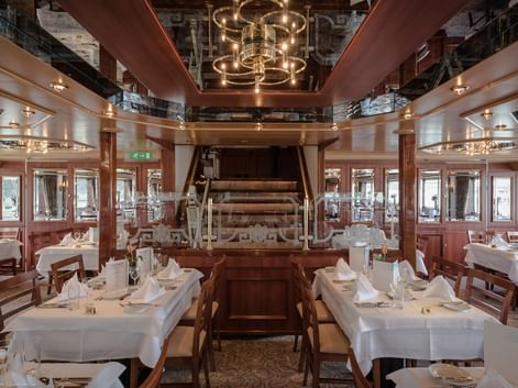 Elegant skibsrestaurant med hvide duge, træstole og historisk skovlhjul som midtpunkt. Spejlloft og varm belysning.