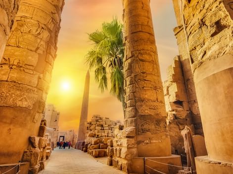 Massive stensøjler med hieroglyfiske udskæringer ved Karnak-templet i Egypten, oplyst af gyldent solnedgangslys med palmer.