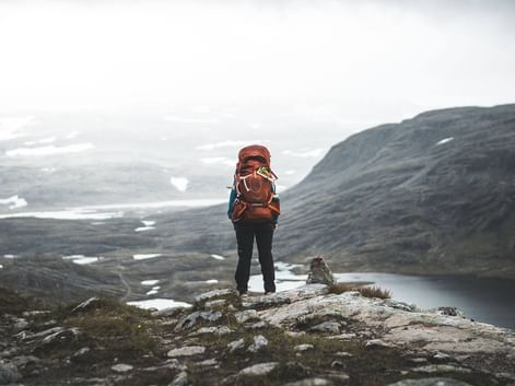 Vandrer med orange rygsæk på klippeformation i Skarvheimen, Norge, med udsigt over bjergplateau med søer og snefelter.