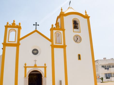 Hvid portugisisk kirke med gule detaljer, to klokketårne og et ur. Kirken har et kors på toppen i Burgau, Lagos.