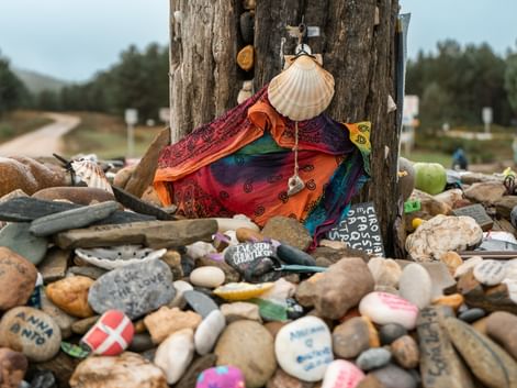 Trestolpe med pilegrimsskjell og fargerikt stoff omgitt av hundrevis av malte steiner etterlatt av Camino-pilgrimer.