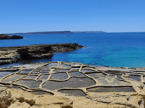 Traditionelle saltpander udskåret i kystklipperne på Gozo, Malta, med turkisblåt Middelhav og fjerne klipper under klar blå himmel.