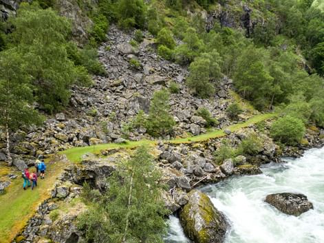 To vandrere i farverige jakker på den smalle Kongevegen-sti ved en rivende turkis flod i en klippefyldt norsk kløft med grøn vegetation.