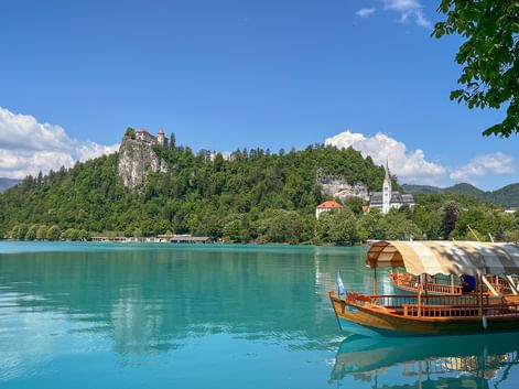 Traditionella träbåtar på det turkosa Bledsjön med Bled slott på en skogbeklädd klippa och ett vitt kyrktorn synligt.