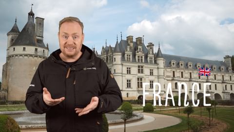 Man in black jacket gesturing in front of French château with towers. Text 'FRANCE' with British flag visible on right side.