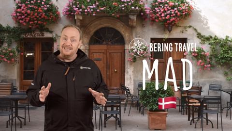 Mand i sort jakke gestikulerer foran blomsterdekoreret bygning med udendørs siddepladser. Tekst viser 'BERING TRAVEL MAD' med dansk flag.