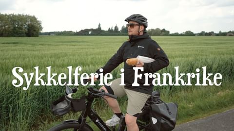 Syklist med hjelm og solbriller står med elsykkel på landevei ved siden av grønne åkrer. Teksten 'Sykkelferie i Frankrike' lagt over bildet.