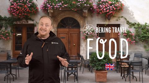 YouTube cover showing a man in black jacket gesturing in front of a rustic building with flower-decorated balconies. Text reads 'FOOD' with UK flag.