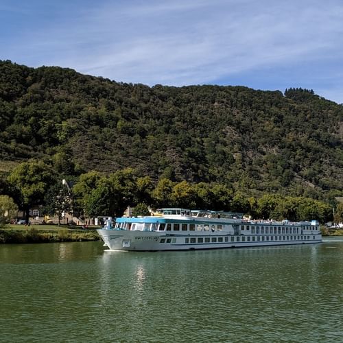 Hvidt flodkrydstogtskib Switzerland sejlende på roligt grønt vand med skovklædte bakker og lille landsby i baggrunden under blå himmel.