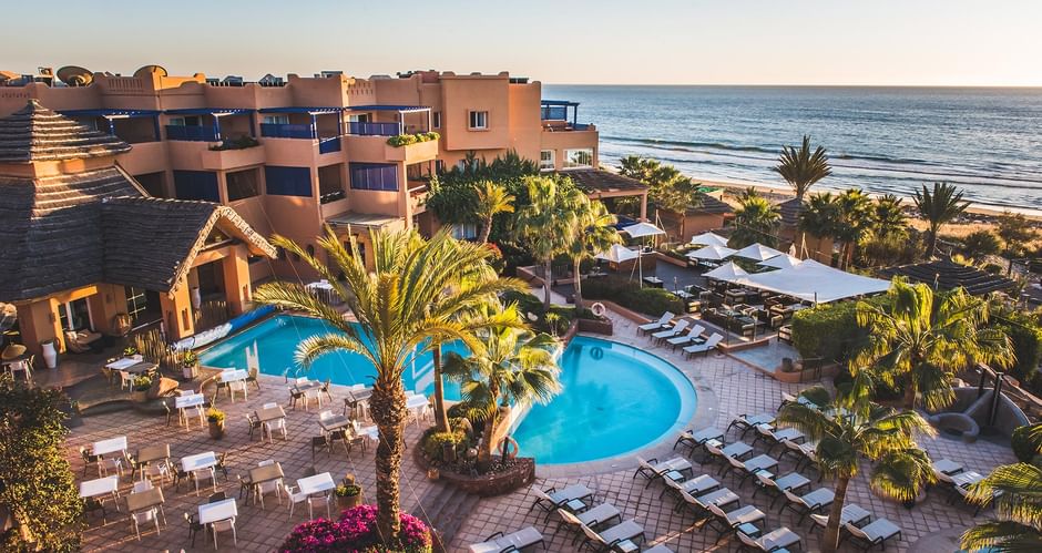 Aerial view of Paradise Plage resort in Agadir with curved swimming pools, palm trees, sun loungers, and beachfront location at sunset.