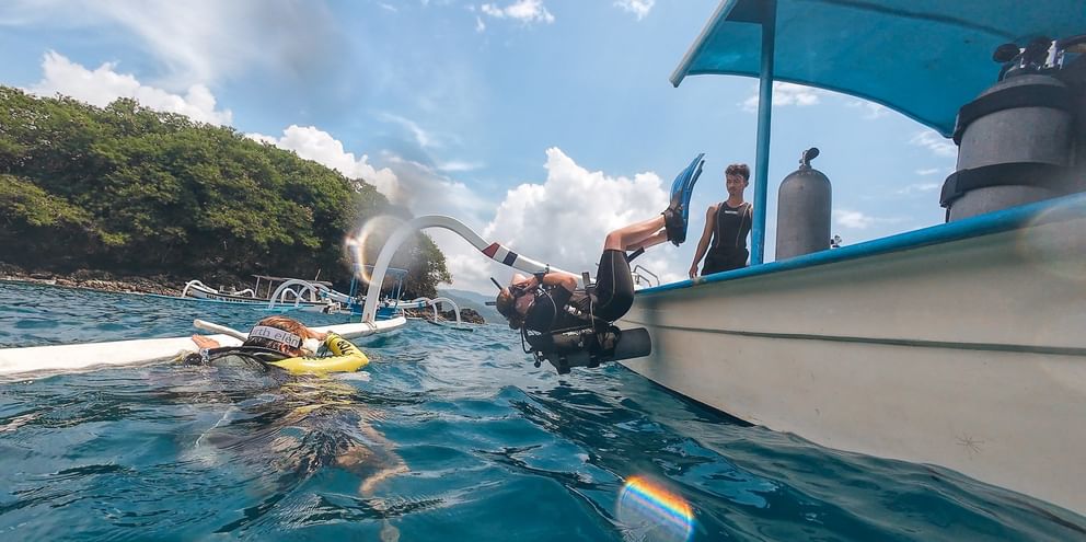 Dykker i våtdrakt går i turkist vann fra hvit dykkerbåt på Bali. Dykkerflasker synlige på båten, grønn kystlinje og blå himmel i bakgrunnen.