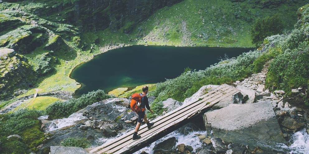Vandrer med orange rygsæk krydser træbro over bjergbæk i Aurlandsdalen, med mørk bjergsø og grønne skråninger nedenfor.