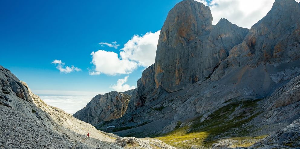 Dramatiske kalkstenstinder ved Picu Urriellu i Picos de Europa med vandrer på klippefyldt skråning. Grøn dal og blå himmel med hvide skyer.