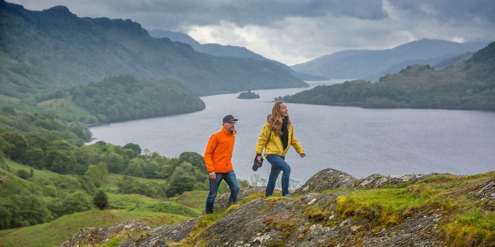 Fotturister på West Highland Way ved innsjø To fotturister i fargerike jakker står på mosegrodd stein med utsikt over en skotsk innsjø omgitt av grønne åser og fjell.