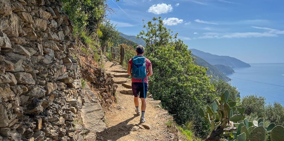Hiker with blue backpack walking on stone path along Ligurian coast. Stone wall on left, Mediterranean Sea and mountains visible on right.