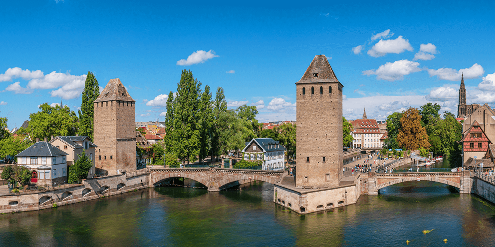 Middelalderlige stentårne flankerer en bro over en kanal i Strasbourg. Historiske bygninger og kirketårne synlige i baggrunden under blå himmel.