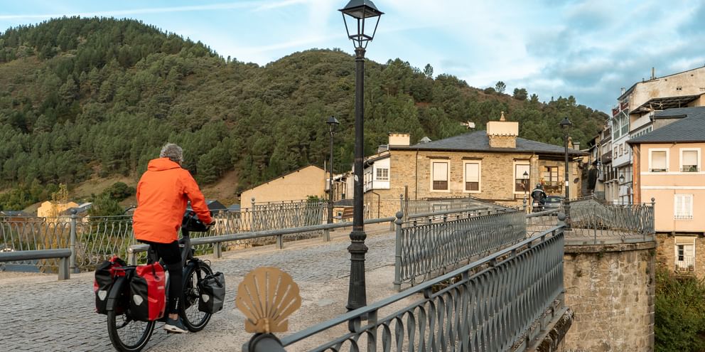 Cyklist i orange jakke med lastet turistcykel krydser stenbro med udsmykkede rækværk. Camino-muslingesymbol synligt på brogelænder.