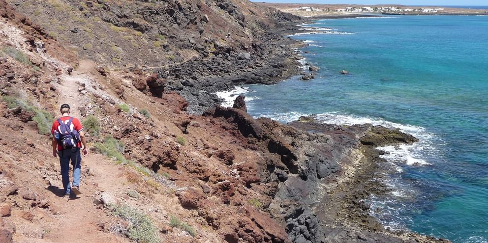 Hiker walking on volcanic rocky coastline of La Graciosa with turquoise ocean waters and distant white buildings visible on the horizon.