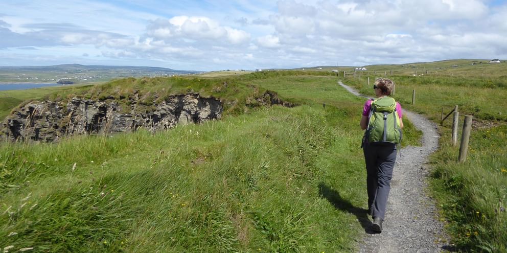 Vandrer med rygsæk går på grusvej langs stenmur i irsk landskab. Grønne marker, fjerne bakker og skyet himmel synlig.