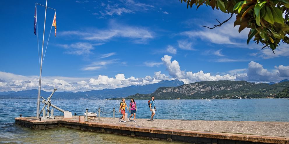 Lake Garda wooden pier with hikers Three hikers on wooden pier at Lake Garda with flagpole, mountains in background under blue sky with white clouds.