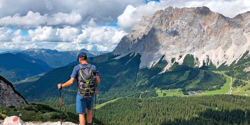 Vandrer med rygsæk og vandrestave ser på dramatiske Zugspitze toppe, grønne dale og skove under overskyet himmel.