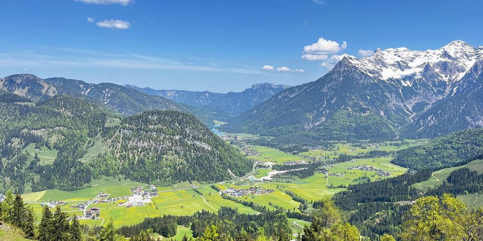 Panoramic view of a green Austrian alpine valley with villages, surrounded by forested mountains and snow-capped peaks under blue sky.