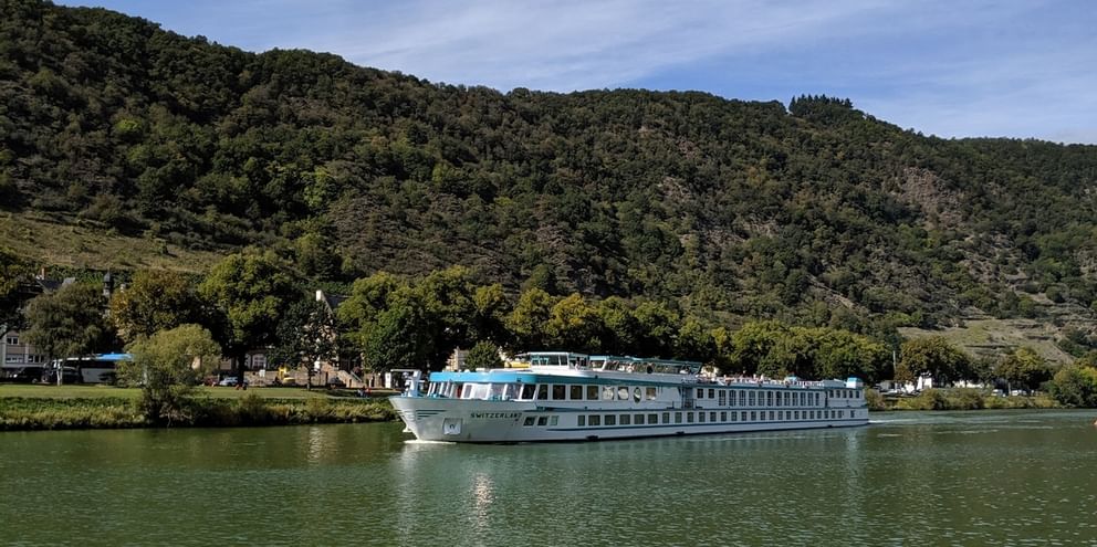 Hvidt flodkrydstogtskib Switzerland sejlende på roligt grønt vand med skovklædte bakker og lille landsby i baggrunden under blå himmel.
