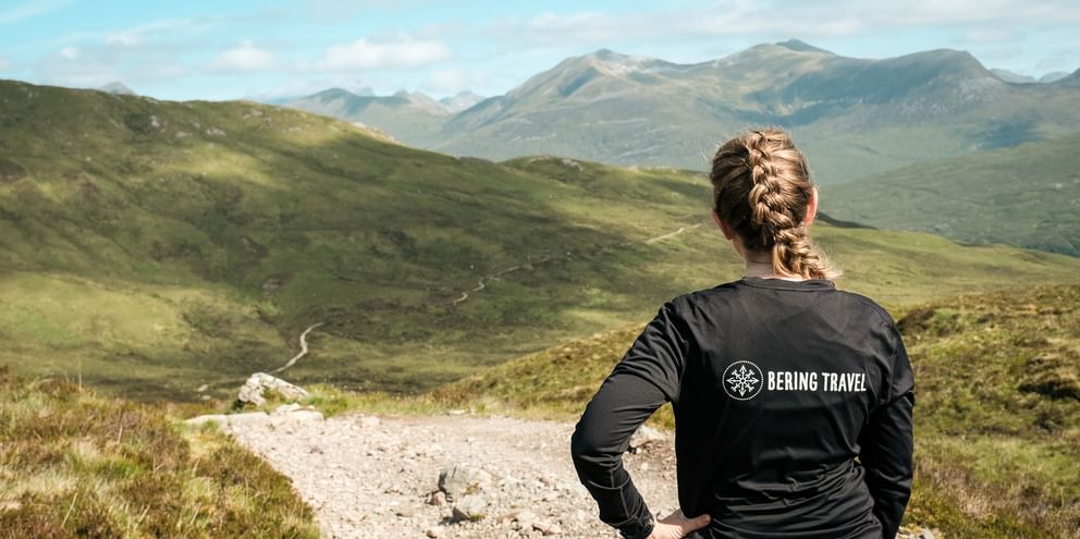 Kvinde med flettet hår i Bering Travel jakke står på sti med udsigt over grønne skotske højlande med bjerge under blå himmel.