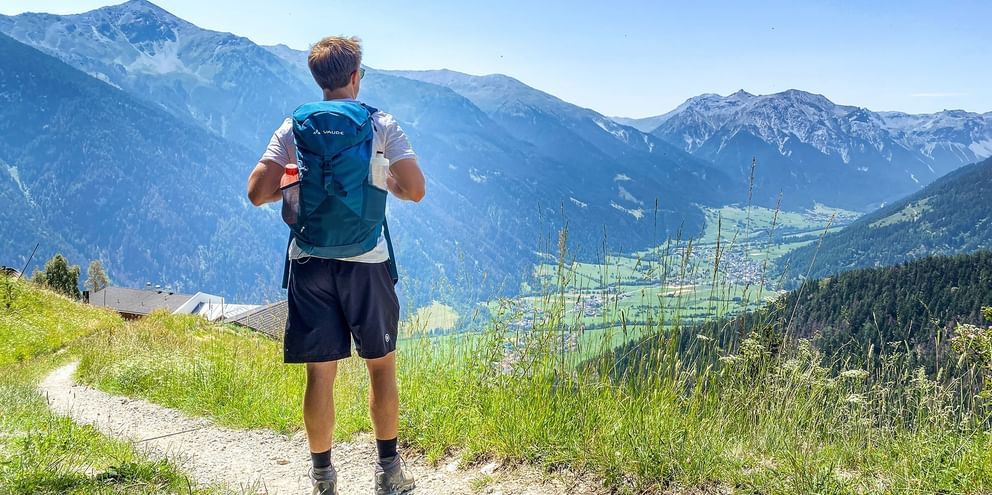 Vandrare med blå ryggsäck på bergsled med utsikt över Alpdal med gröna ängar och snötäckta toppar vid Garmisch.