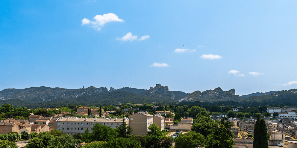 Panoramaudsigt over Saint-Rémy-de-Provence med terracottatage og cypresser, med de klippefyldte Alpilles-bjerge i baggrunden under blå himmel.