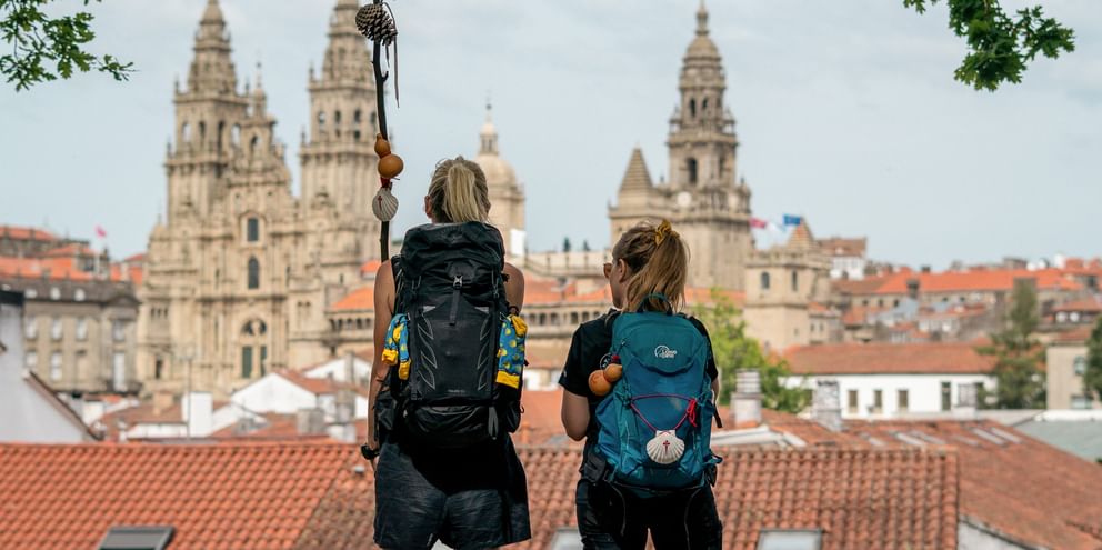 Two hikers with backpacks viewing Santiago de Compostela Cathedral from a rooftop. The cathedral's towers rise above red-tiled roofs.