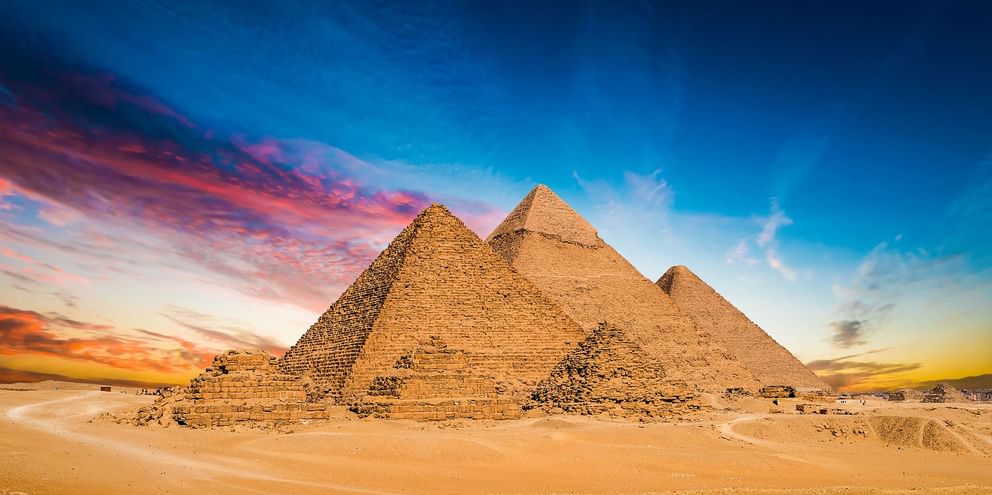Tre gamle pyramider i Giza i den egyptiske ørken under en dramatisk solnedgangshimmel med orange og blå skyer.