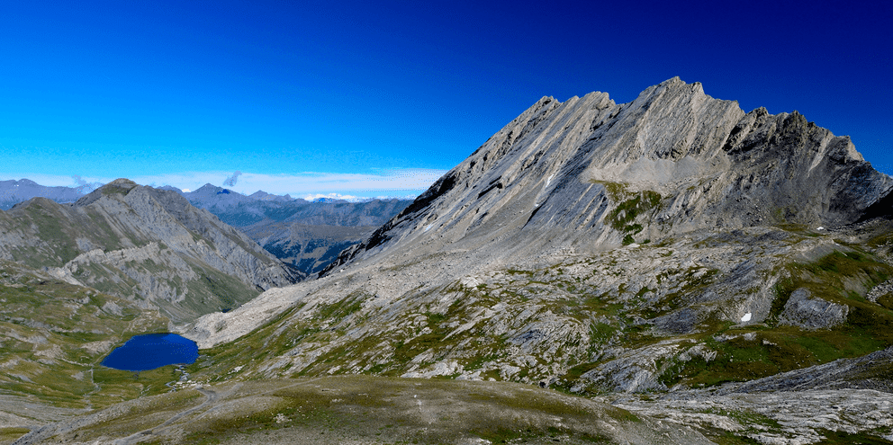 Klippefyldt bjergtop med lagdelte grålige skråninger over en lille blå alpesø i Queyras Naturpark, Frankrig, under klar blå himmel.