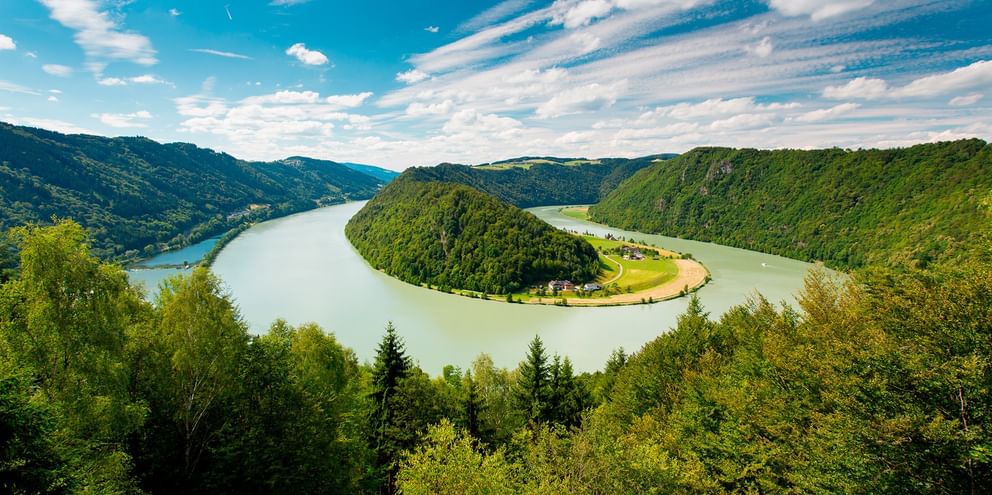 Luftfoto af Donau, der danner en hestesko-sløjfe omkring en skovklædt bakke i Østrig, med grønne skove og blå himmel.
