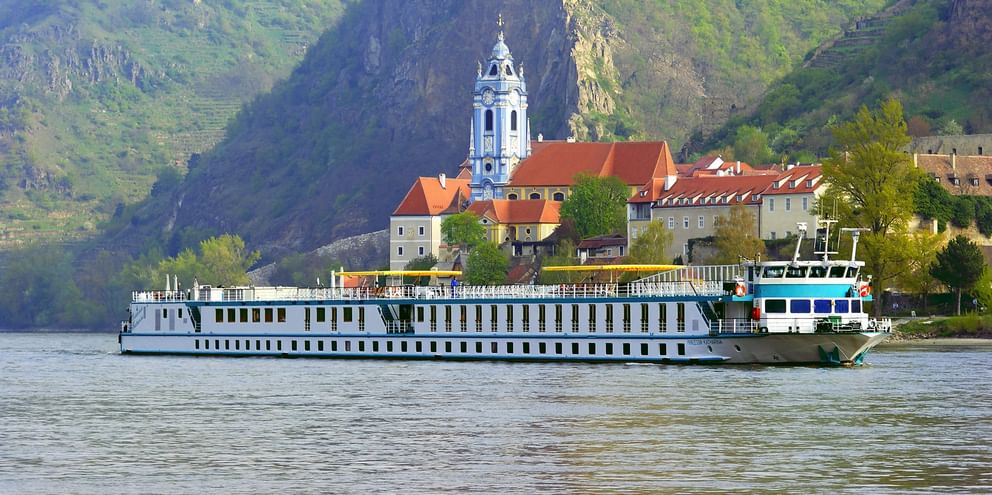 White river cruise ship sailing on the Danube with historic town featuring baroque church tower and red-roofed buildings against green hills.