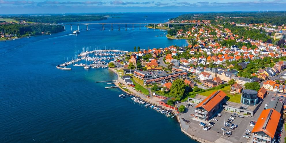 Flygbild över Svendborg, Danmark som visar småbåtshamnen, färgglada hus, strandbyggnader och en bro över det blå vattnet.