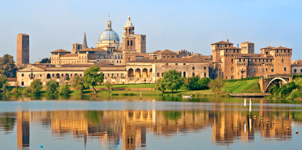 Historisk Mantova skyline med domkuppel og klokketårn spejlet i roligt Po-vand, omgivet af grøn vegetation under blå himmel.