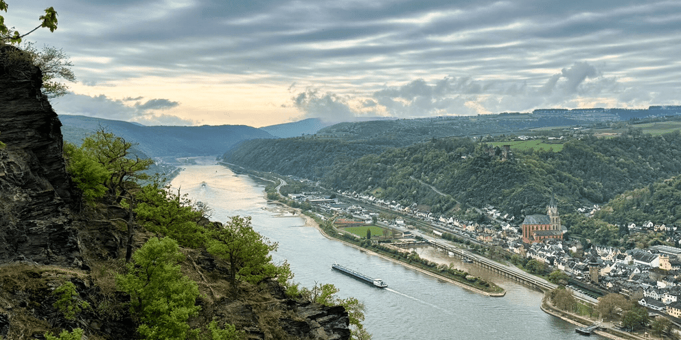 Panoramaudsigt over Rhinen, der slynger sig gennem dalen med fragtskib, byer på begge bredder, grønne bakker og dramatisk skyet himmel.