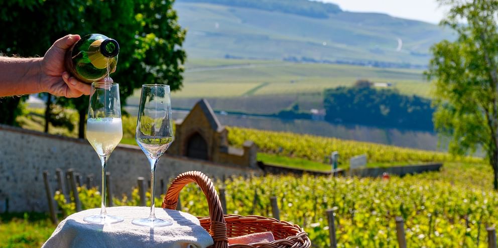 Hånd heller champagne i glass på hvitt bord med kurv, med utsikt over Champagne-vingårder og bølgende åser.